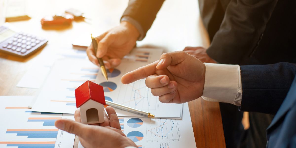 Two businesspeople discussing charts and graphs on a table while holding a small house model, representing a real estate strategy meeting.